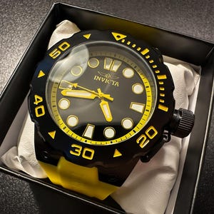 May include: A black and yellow Invicta watch with a black bezel and yellow detailing. The watch has a black face with yellow hands and markers. The watch is presented in a black box with a white cushion.