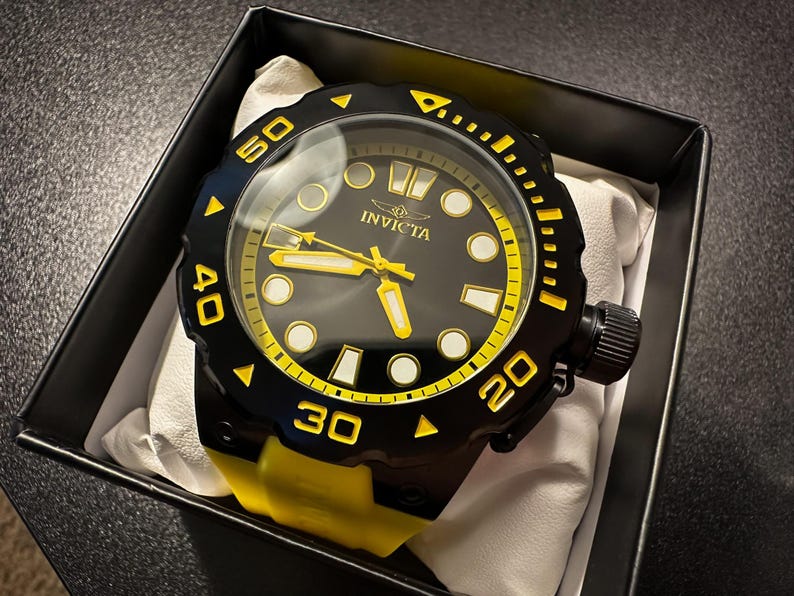 May include: A black and yellow Invicta watch with a black bezel and yellow detailing. The watch has a black face with yellow hands and markers. The watch is presented in a black box with a white cushion.