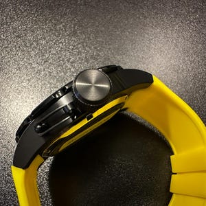 May include: A black wristwatch with a yellow strap. The watch features a black face and a silver crown. The yellow strap is made of a flexible material.