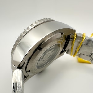 May include: A close-up of a silver wristwatch with a textured bezel. The watch has a metallic strap and a clear back, showing the inner workings. A yellow tag is attached to the strap. The watch is new and unworn.