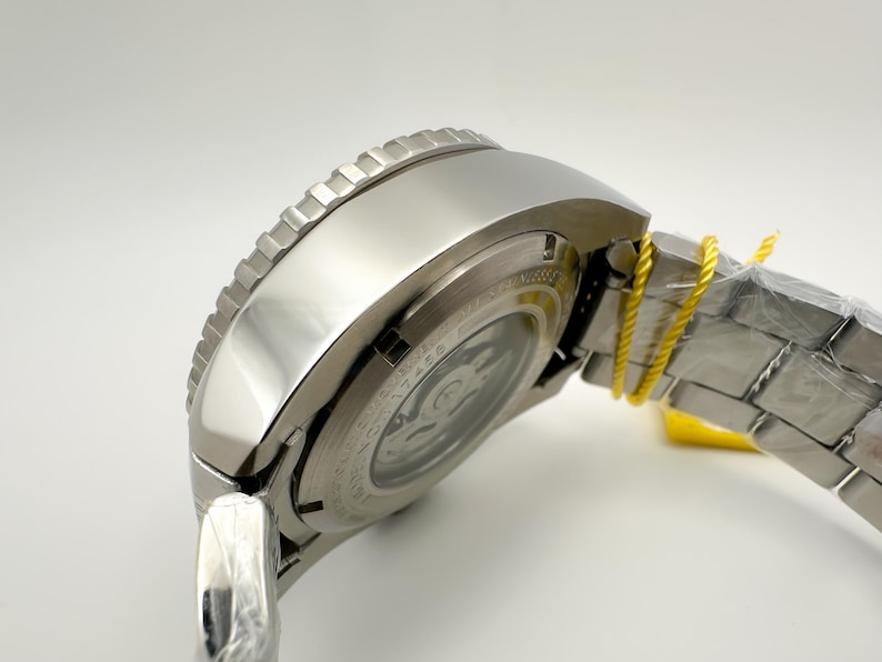May include: A close-up of a silver wristwatch with a textured bezel. The watch has a metallic strap and a clear back, showing the inner workings. A yellow tag is attached to the strap. The watch is new and unworn.