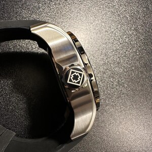 May include: A side view of a wristwatch featuring a brushed silver metal case and a black rubber strap. The crown is black with a white geometric emblem consisting of a square outline and a central cross shape, matching the watch's design.
