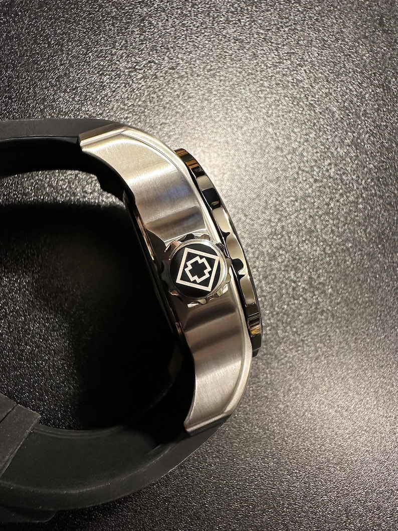 May include: A side view of a wristwatch featuring a brushed silver metal case and a black rubber strap. The crown is black with a white geometric emblem consisting of a square outline and a central cross shape, matching the watch's design.