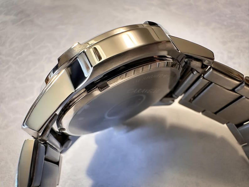 May include: A close-up of a silver wristwatch with a stainless steel band. The watch face is visible, and the band is connected to the watch case. The watch has a shiny, polished finish, reflecting the light. The watch is a classic style.