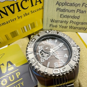 May include: A silver Invicta watch with a brown face and visible inner workings. The watch has a textured bezel and a stainless steel strap. The image includes yellow and grey cards with text, including "Invicta" and "Five Year Warranty."