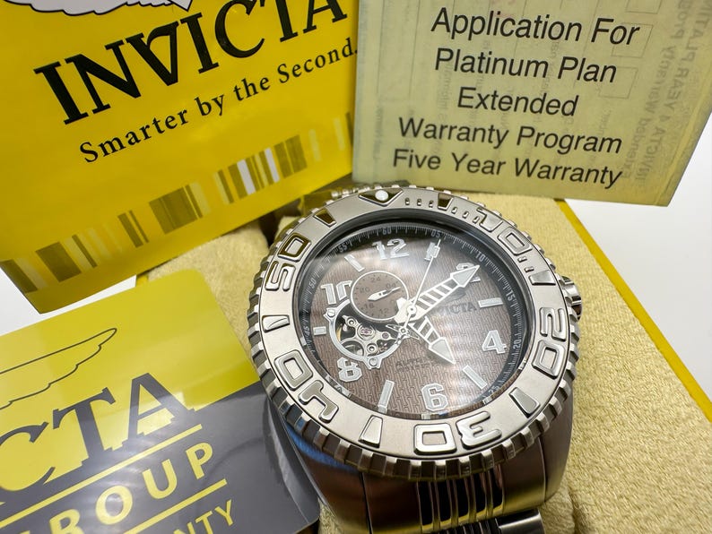 May include: A silver Invicta watch with a brown face and visible inner workings. The watch has a textured bezel and a stainless steel strap. The image includes yellow and grey cards with text, including "Invicta" and "Five Year Warranty."