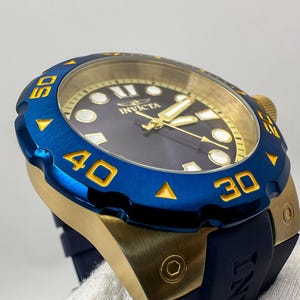 May include: A close-up of a blue and gold Invicta watch. The watch has a blue bezel with gold numerals and a gold-toned case. The watch face is dark blue with gold hands and markers. The watch band is dark blue.