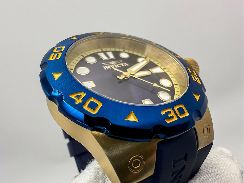 May include: A close-up of a blue and gold Invicta watch. The watch has a blue bezel with gold numerals and a gold-toned case. The watch face is dark blue with gold hands and markers. The watch band is dark blue.