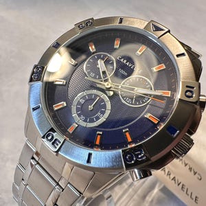 May include: Silver-toned Caravelle watch with a blue face and silver band. The watch features three sub-dials, orange accents, and a 100m water resistance rating. The watch is new with a tag attached.