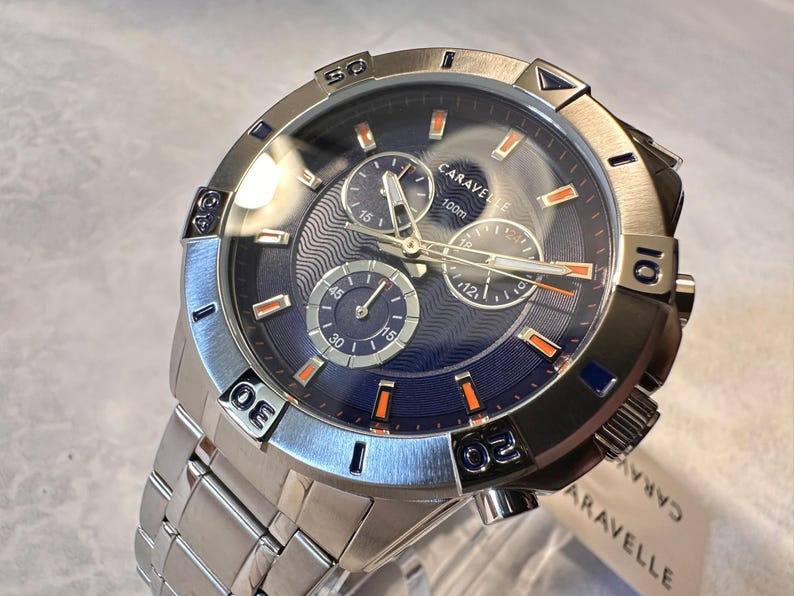 May include: Silver-toned Caravelle watch with a blue face and silver band. The watch features three sub-dials, orange accents, and a 100m water resistance rating. The watch is new with a tag attached.