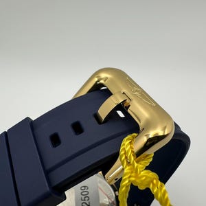 May include: Close-up of a gold-colored watch clasp and a navy blue watch band. The clasp is rectangular with a curved design and a logo. A yellow tag is attached to the watch. The watch band has two square holes.