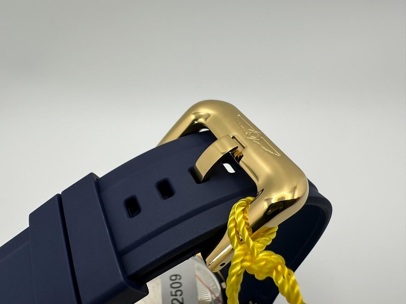 May include: Close-up of a gold-colored watch clasp and a navy blue watch band. The clasp is rectangular with a curved design and a logo. A yellow tag is attached to the watch. The watch band has two square holes.