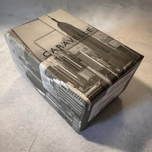 May include: A rectangular box wrapped in paper featuring a black and white image of the New York City skyline, including the Empire State Building. The word "CARAVELLE" is printed on the box.