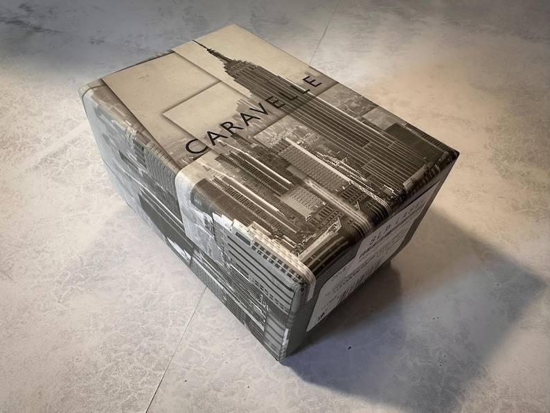 May include: A rectangular box wrapped in paper featuring a black and white image of the New York City skyline, including the Empire State Building. The word "CARAVELLE" is printed on the box.