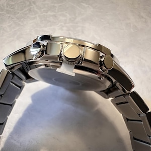 May include: A stainless steel wristwatch with a silver band and a round face. The watch has three buttons on the side and a polished finish. The watch band is made of rectangular links.