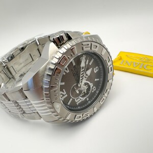 May include: A silver Invicta watch with a dark grey face and a stainless steel strap. The watch has a textured bezel and is next to a yellow Invicta tag.