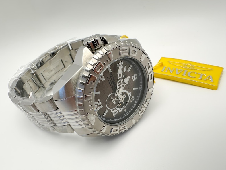 May include: A silver Invicta watch with a dark grey face and a stainless steel strap. The watch has a textured bezel and is next to a yellow Invicta tag.