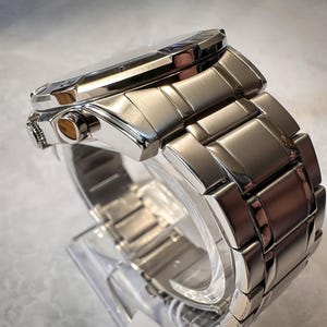 May include: A close-up side view of a silver-toned wristwatch. The watch features a rectangular face and a linked metal band. The watch is displayed on a clear plastic stand, showcasing its polished finish and intricate design.