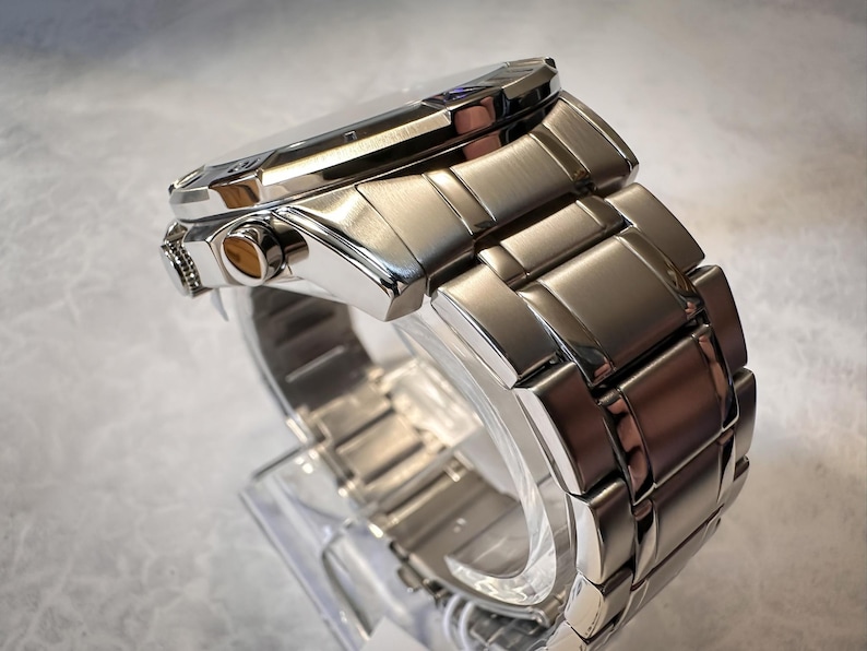 May include: A close-up side view of a silver-toned wristwatch. The watch features a rectangular face and a linked metal band. The watch is displayed on a clear plastic stand, showcasing its polished finish and intricate design.