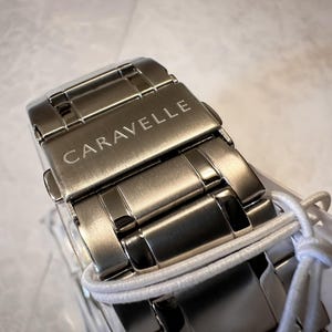 May include: Close-up of a silver-toned Caravelle watch clasp. The watch band is made of interlocking metal links. The word "CARAVELLE" is engraved on the clasp. A white string is wrapped around the watch band.
