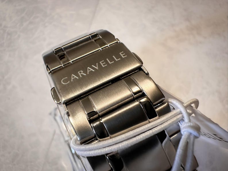 May include: Close-up of a silver-toned Caravelle watch clasp. The watch band is made of interlocking metal links. The word "CARAVELLE" is engraved on the clasp. A white string is wrapped around the watch band.