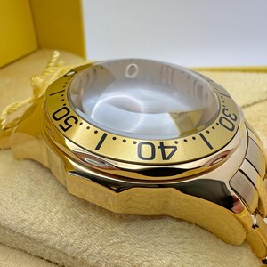 May include: A gold-toned wristwatch with a clear, domed crystal. The watch face features black numerals and markers against a gold bezel. The watch band is a combination of gold and silver-toned metal. The watch is presented in a yellow box.