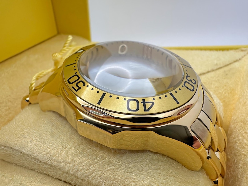 May include: A gold-toned wristwatch with a clear, domed crystal. The watch face features black numerals and markers against a gold bezel. The watch band is a combination of gold and silver-toned metal. The watch is presented in a yellow box.