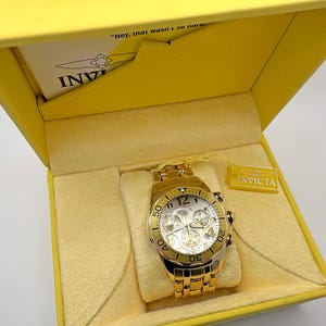 May include: Gold-toned Invicta chronograph watch with a white dial, displayed in a yellow box. The watch features multiple sub-dials and a date display. The box has a yellow interior and the Invicta logo is visible.