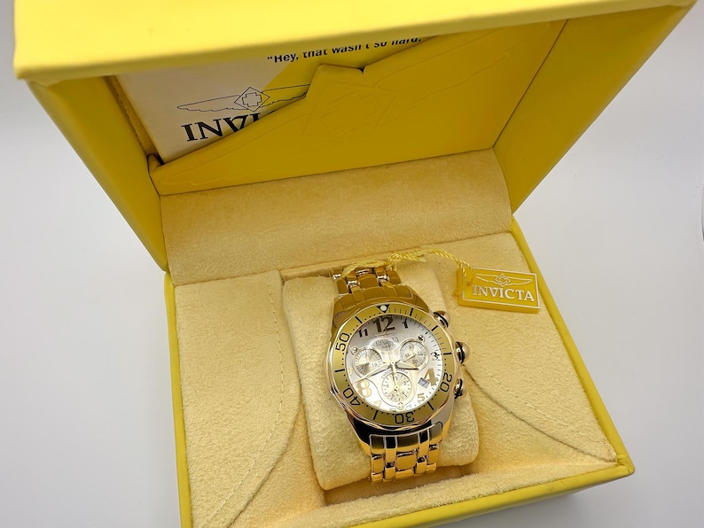 May include: Gold-toned Invicta chronograph watch with a white dial, displayed in a yellow box. The watch features multiple sub-dials and a date display. The box has a yellow interior and the Invicta logo is visible.