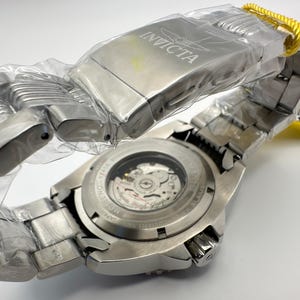 May include: A new Invicta stainless steel watch, still in its protective plastic. The watch features a silver band and a visible mechanical movement on the back. The watch has a yellow tag attached.