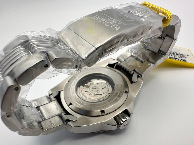 May include: A new Invicta stainless steel watch, still in its protective plastic. The watch features a silver band and a visible mechanical movement on the back. The watch has a yellow tag attached.