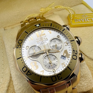 May include: A gold-toned Invicta chronograph watch with a white dial. The watch features gold numerals, hands, and sub-dials. The watch is water-resistant to 200 meters (660 feet) and has a gold link bracelet.