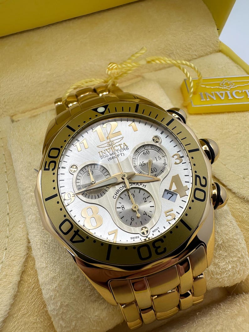 May include: A gold-toned Invicta chronograph watch with a white dial. The watch features gold numerals, hands, and sub-dials. The watch is water-resistant to 200 meters (660 feet) and has a gold link bracelet.