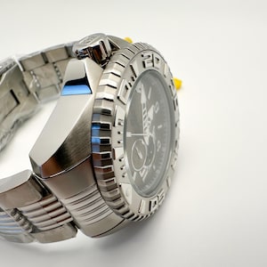 May include: A silver wristwatch with a black dial and a textured bezel. The watch has a stainless steel strap and a blue accent on the side. The watch is new and still has protective plastic on the strap.
