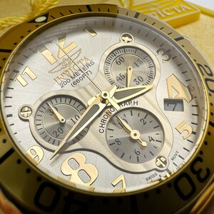 May include: Gold-toned Invicta chronograph watch with a silver dial. Features include three sub-dials, luminous hands, and a date window. The watch is water-resistant to 200 meters (660 feet).