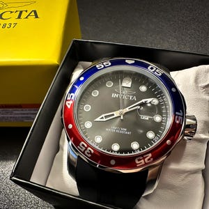 May include: An Invicta wristwatch with a black dial, silver-tone hands, and circular hour markers. The bezel features a red and blue color scheme with white numerals. The watch has a black strap and is presented in a black box with a yellow lid.