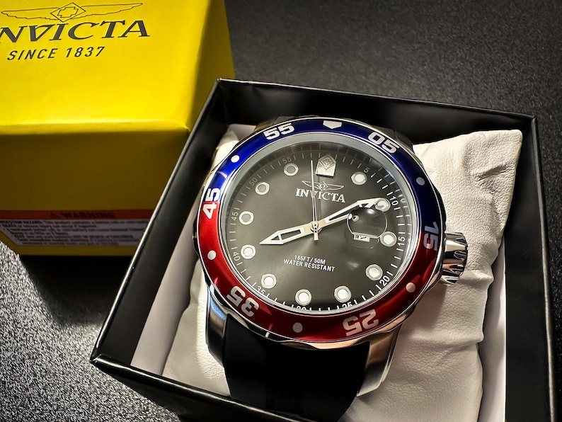 May include: An Invicta wristwatch with a black dial, silver-tone hands, and circular hour markers. The bezel features a red and blue color scheme with white numerals. The watch has a black strap and is presented in a black box with a yellow lid.