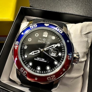 May include: An Invicta wristwatch with a black dial, luminous markers, and a date window at the three o'clock position. The bezel features a blue and red color scheme with white numerals. The watch has a black strap and is water resistant to 165FT.