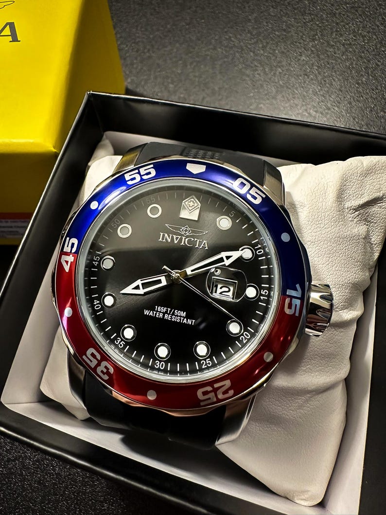 May include: An Invicta wristwatch with a black dial, luminous markers, and a date window at the three o'clock position. The bezel features a blue and red color scheme with white numerals. The watch has a black strap and is water resistant to 165FT.