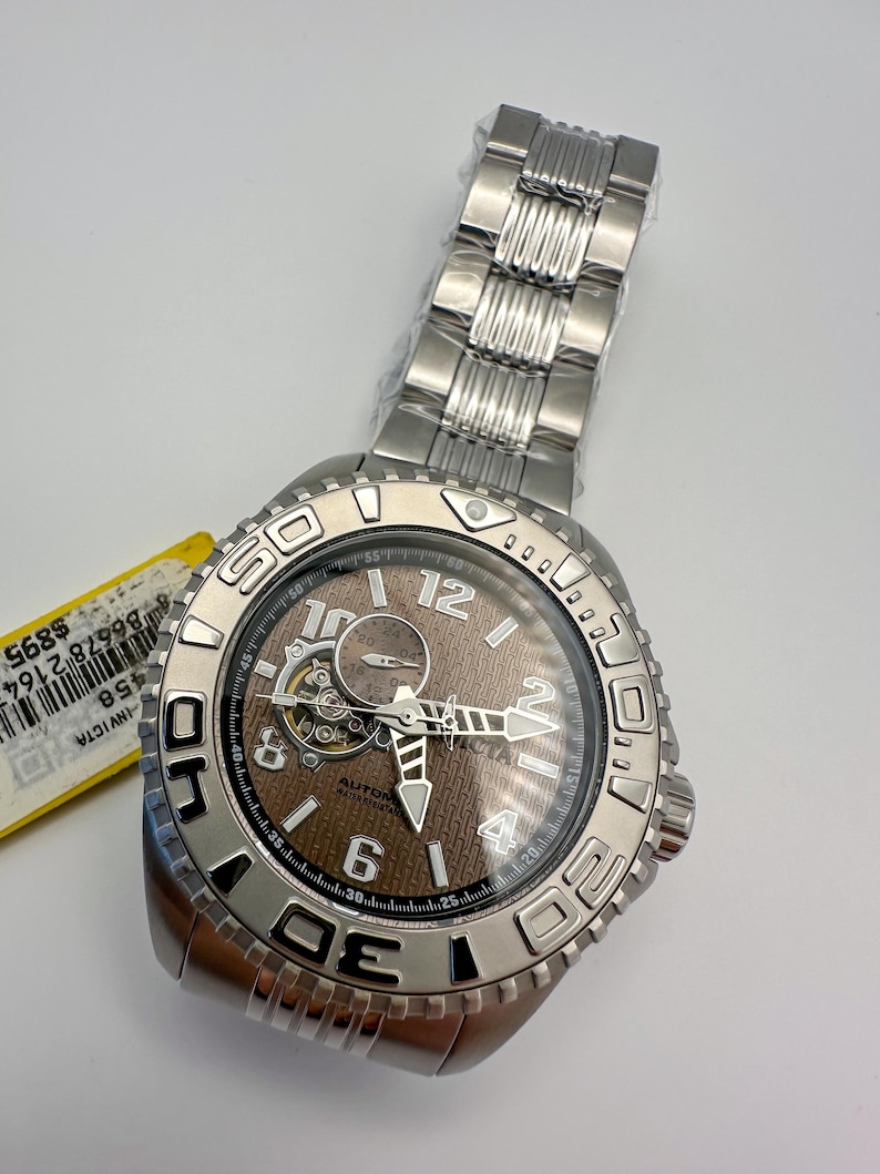 May include: A silver Invicta automatic watch with a brown face and a stainless steel strap. The watch has a visible mechanism and is marked with the numbers 6, 12, and 4. The watch is new and still has the original packaging.