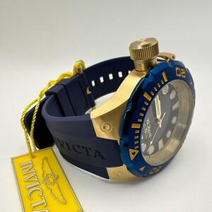 May include: A gold-toned watch with a blue bezel and a dark blue band. The watch face is black with gold accents. A yellow tag with the brand name is attached to the watch.