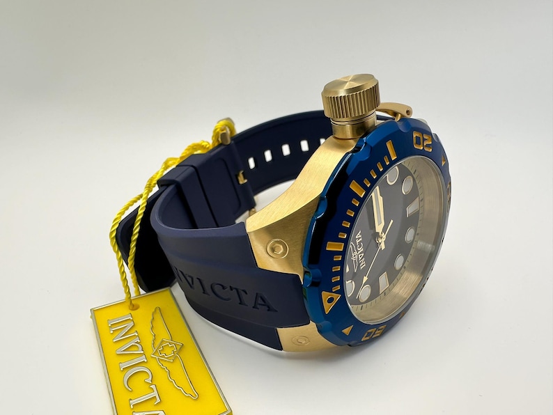 May include: A gold-toned watch with a blue bezel and a dark blue band. The watch face is black with gold accents. A yellow tag with the brand name is attached to the watch.