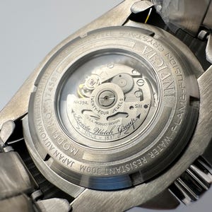 May include: A close-up of the back of a silver Invicta Fusion watch. The watch has a clear crystal back, showcasing the automatic movement's inner workings. Text includes "Invicta", "Flame-Fusion Crystal", "Water Resistant 300m", and "Japan Automatic Movement".