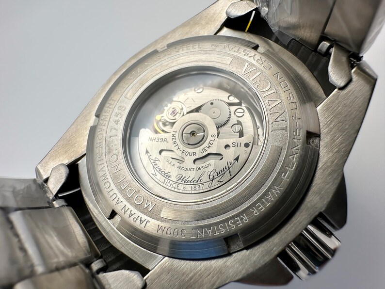 May include: A close-up of the back of a silver Invicta Fusion watch. The watch has a clear crystal back, showcasing the automatic movement's inner workings. Text includes "Invicta", "Flame-Fusion Crystal", "Water Resistant 300m", and "Japan Automatic Movement".