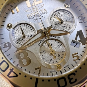 May include: Close-up of a gold and silver Invicta chronograph watch. The watch face features three sub-dials, gold hands, and a date display. The bezel has black markings. The watch is water-resistant up to 200 meters (660 ft).