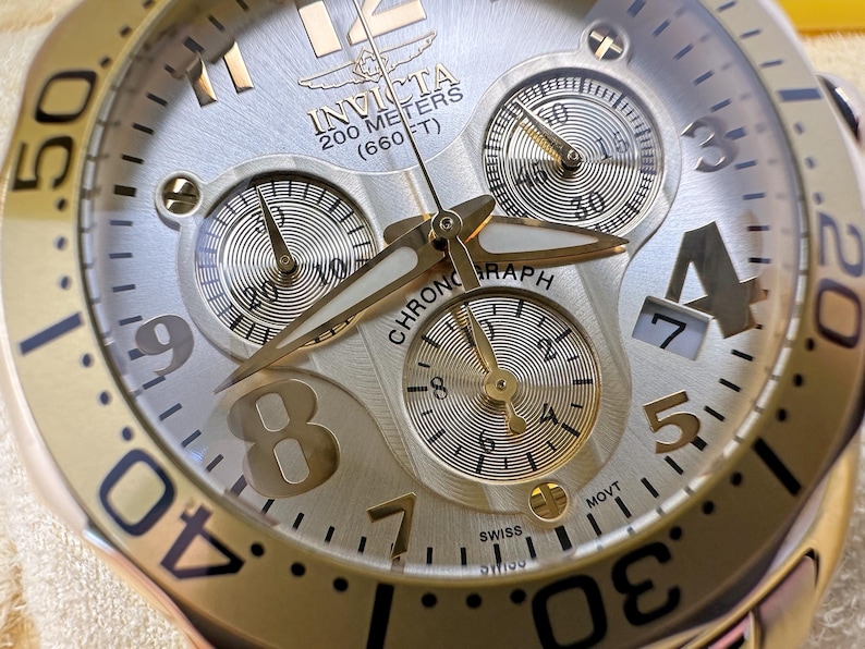May include: Close-up of a gold and silver Invicta chronograph watch. The watch face features three sub-dials, gold hands, and a date display. The bezel has black markings. The watch is water-resistant up to 200 meters (660 ft).