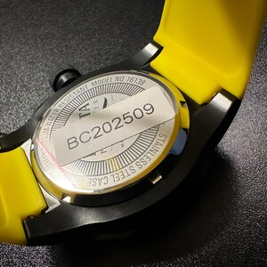 May include: A close-up shot of a black watch with a yellow strap. The watch features a stainless steel case and a black crown. The watch back has a sticker with the text "BC202509". The watch is water resistant to 300m.