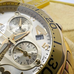 May include: Close-up of a gold-toned Invicta chronograph watch. The watch face is silver with gold accents, featuring multiple sub-dials and the text "200 METERS (660 FT)". The bezel has black numerals. The watch is Swiss made.