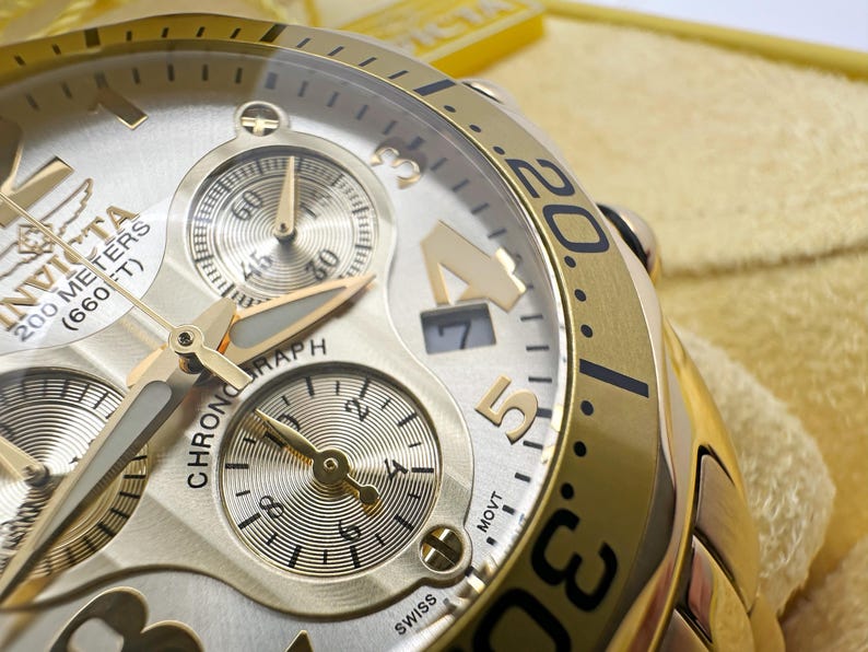 May include: Close-up of a gold-toned Invicta chronograph watch. The watch face is silver with gold accents, featuring multiple sub-dials and the text "200 METERS (660 FT)". The bezel has black numerals. The watch is Swiss made.