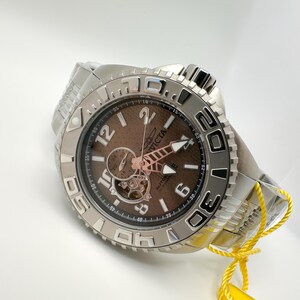May include: Silver Invicta watch with a brown face and visible mechanical movement. The watch has a textured dial, luminous hands, and a stainless steel band. The watch has a rotating bezel with numerals and is new with a yellow tag.
